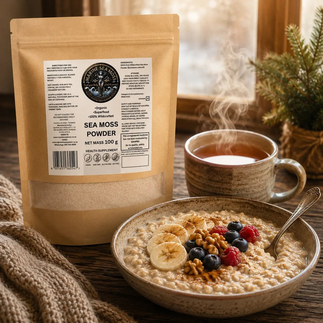 An oatmeal bowl topped with sliced bananas, berries, walnuts, and cinnamon sits next to a steaming mug and a brown paper pouch of "Marine Sea Moss Powder".