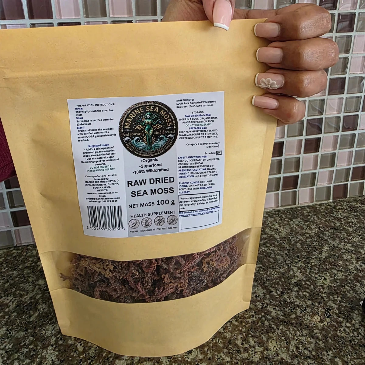 A hand holding a 100g stand-up pouch of Raw Dried Sea Moss against a kitchen countertop and tiled backsplash background. The pouch is a matte kraft paper color with a clear window at the bottom showing the dark brown seaweed inside.